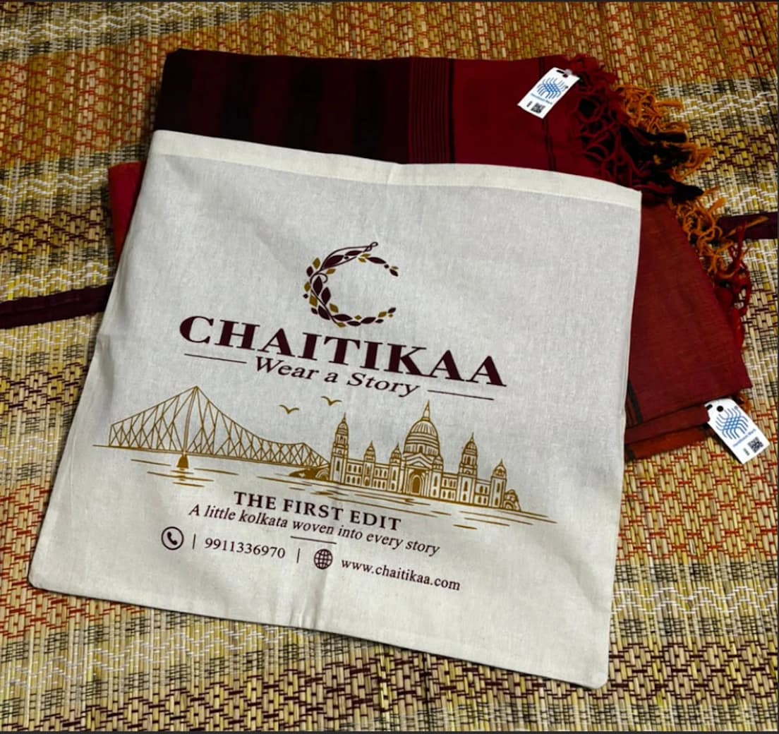 Chaitikaa cotton bag printed with the brand logo and a Kolkata skyline, resting on a maroon handloom saree